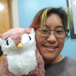 An Asian woman with colorful hair holds an Owlbear plush toy next to her head